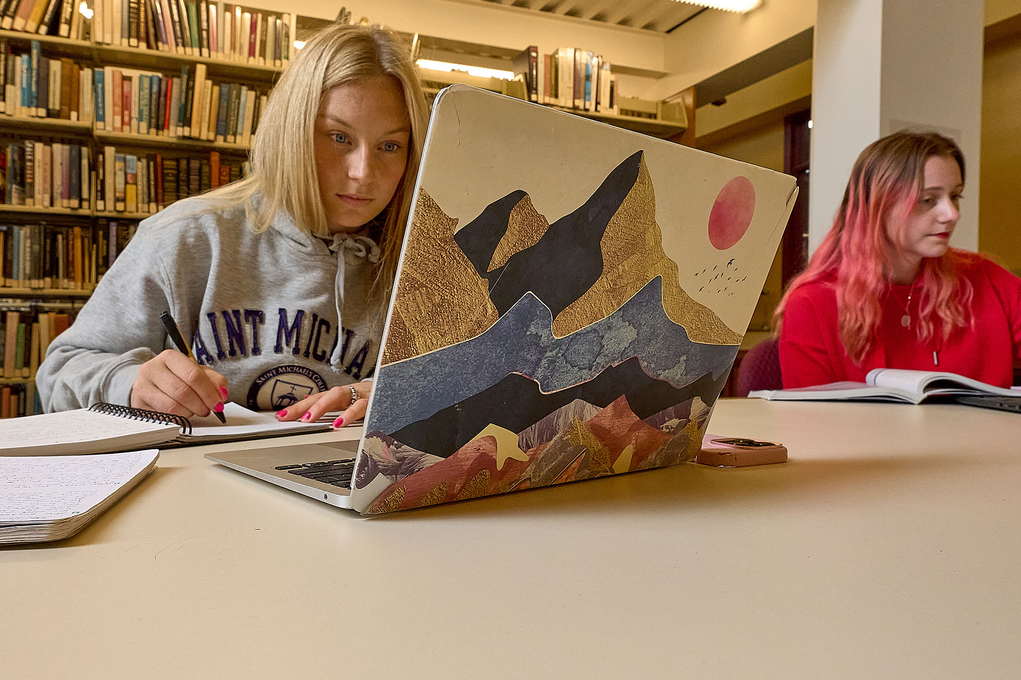 A Saint Michael's College student makes notes in a notebook while looking at her laptop.
