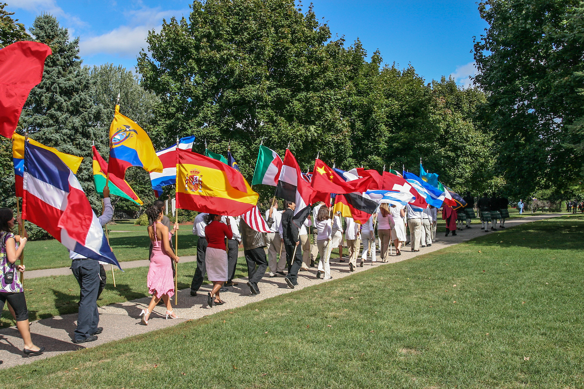 students process though the miffle of campus on a summer day, carrying large international flags.