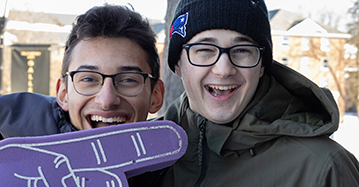 two male students smile at the camera with a large purple foam finger in the bottom corner