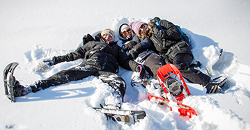 three students lie smiling in snow while wearing snowshoes