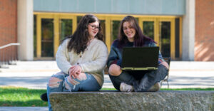 Two female students sitting atop a slab of granite looking at latop and smiling with a set of gold framed entrance doors behind them to a building.