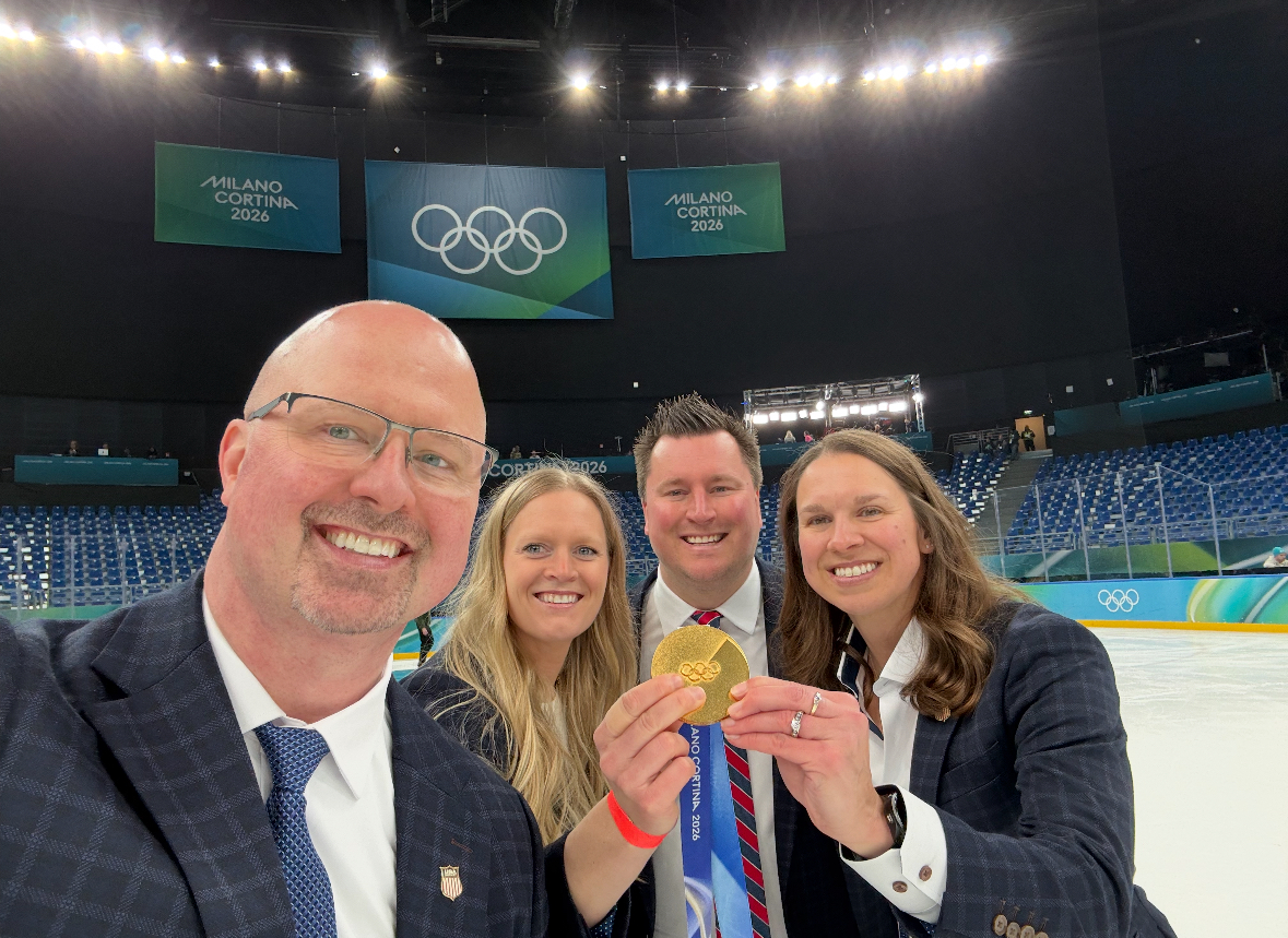 Four people pose in the Olympic hockey arena holding a gold medal.