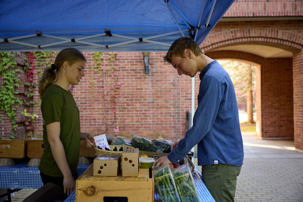 One student works the farm stand, while another looks at items for sale at the table.
