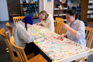 Three people sit at a table and embroider a decorated cloth. 