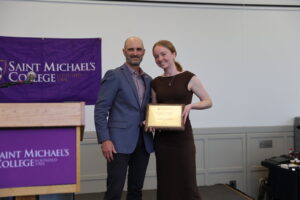 A young woman holds a plaque and poses with a man in a blazer during an awards ceremony.