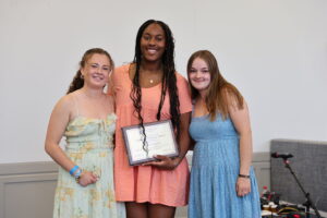Three young women, one with a plaque in hand, pose for a photo during an awards ceremony.