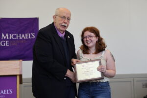 A young woman holds a plaque and poses with an older man in a blazer and tie during an awards ceremony.
