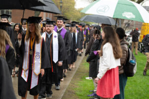 Graduates process in a line toward Commencement, many holding umbrellas, as supporters line the path. 