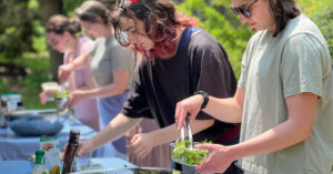 A side profile of a student in a white t-shirt wearing sunglasses filling a salad bowl from contents on a picnic table buffet. A girl in a black shirt next to her is leaning over the table and filling her bowl as well.