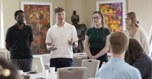 Four students—two male, two female—presenting to a seated audience. They are dressed in casual and smart-casual attire.