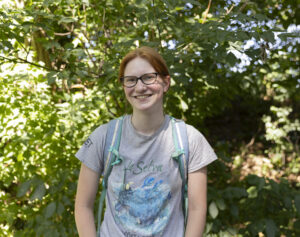 A female student with red hair and glasses smiles for a photo while surrounded by greenery and trees in the Natural Area at Saint Michael's College. 