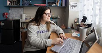 A student with long brown hair and glasses, wearing a Saint Michael's College jersey, smiles at the screen of her open laptop.